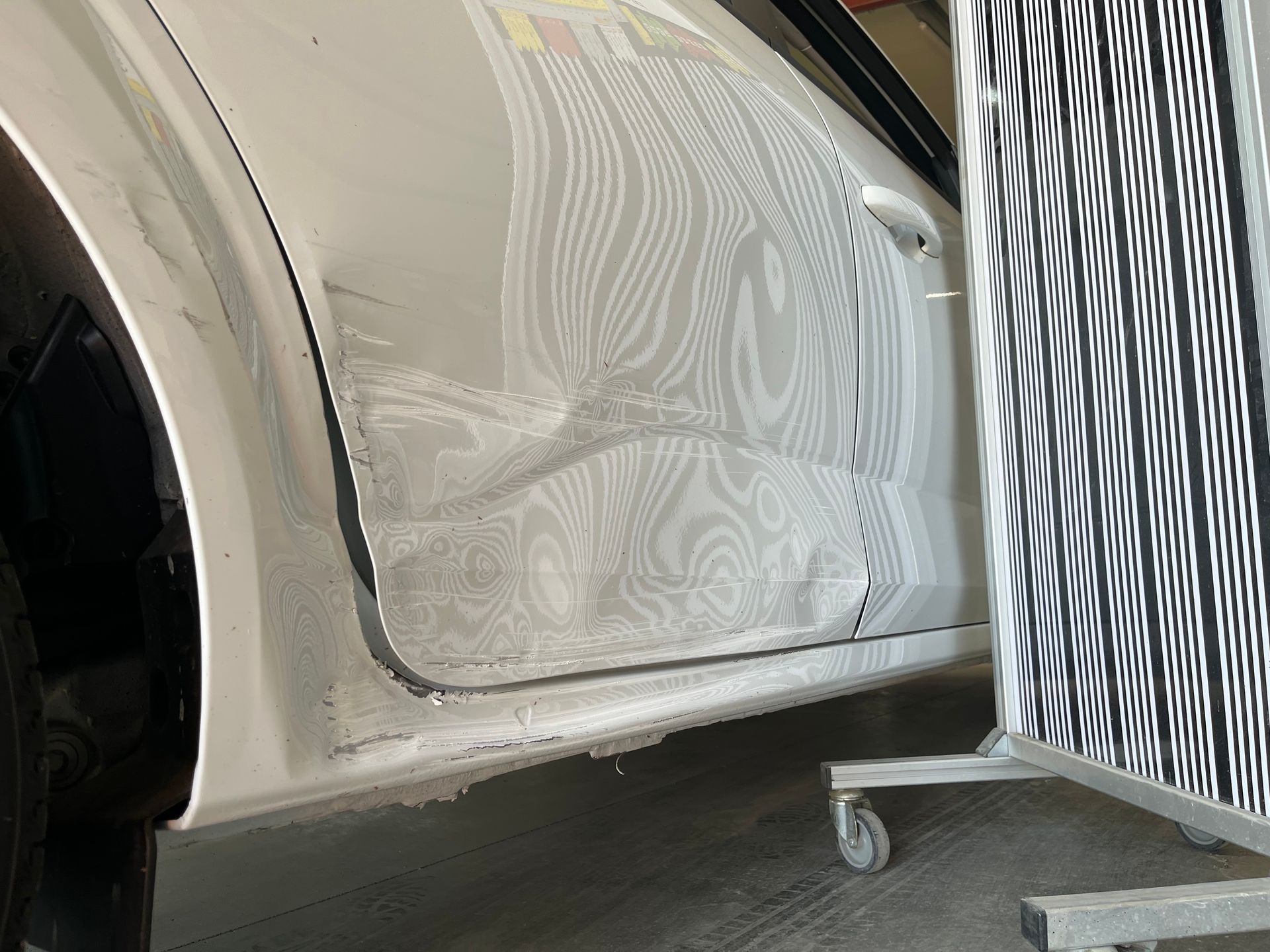Panneau inférieur de portière de voiture avec une bosse et des éraflures, à côté d’un support de réparation rayé