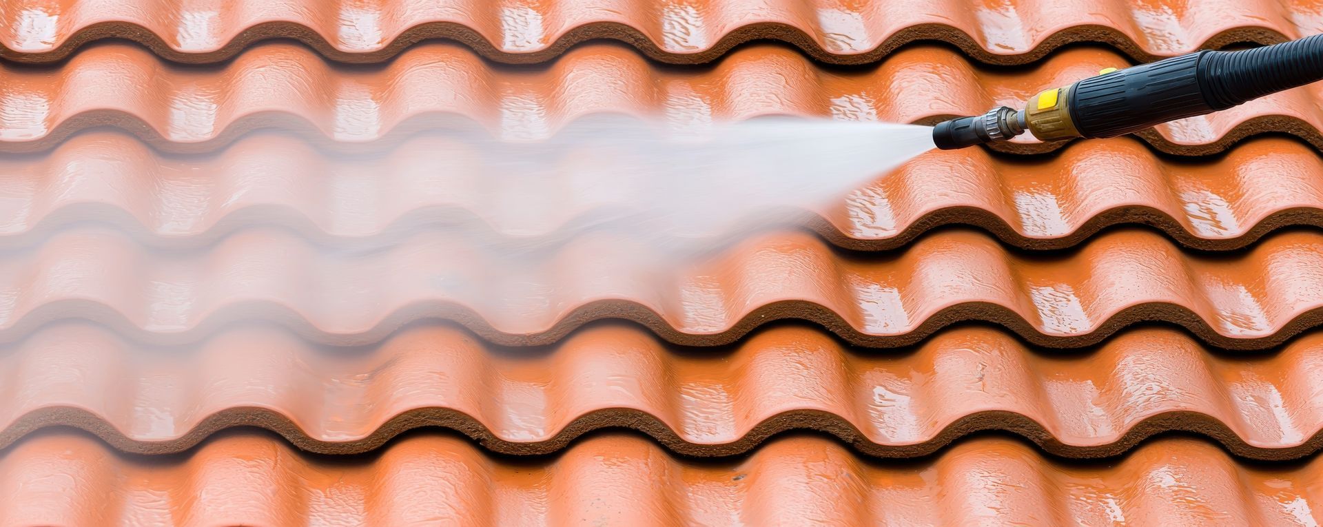 Une personne utilise un nettoyeur haute pression pour nettoyer des tuiles de toit orange à motifs ondulés.