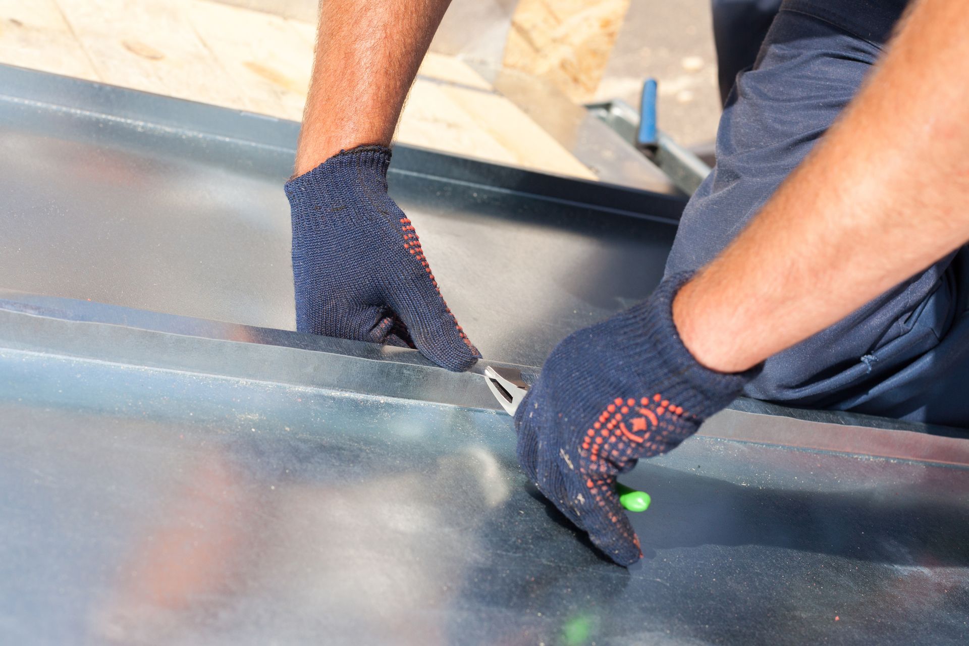 Deux mains gantées utilisent une pince pour sertir le joint de panneaux de toiture métalliques sur une structure en bois.