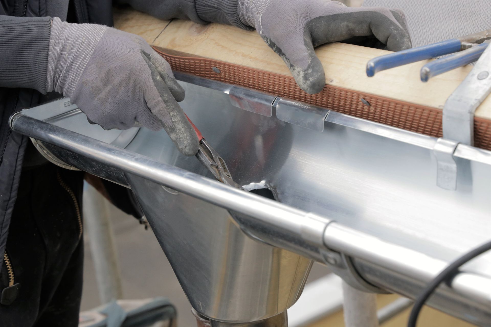 Une personne portant des gants de travail utilise une pince pour installer une sortie de gouttière métallique sur une gouttière de toit.