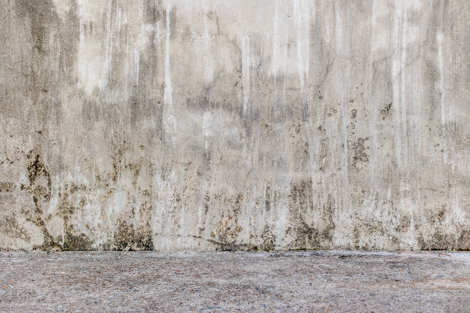 Gros plan sur un mur en béton texturé et patiné, présentant des taches sombres et striées, et une surface au premier plan rugueuse et gravillonnée.