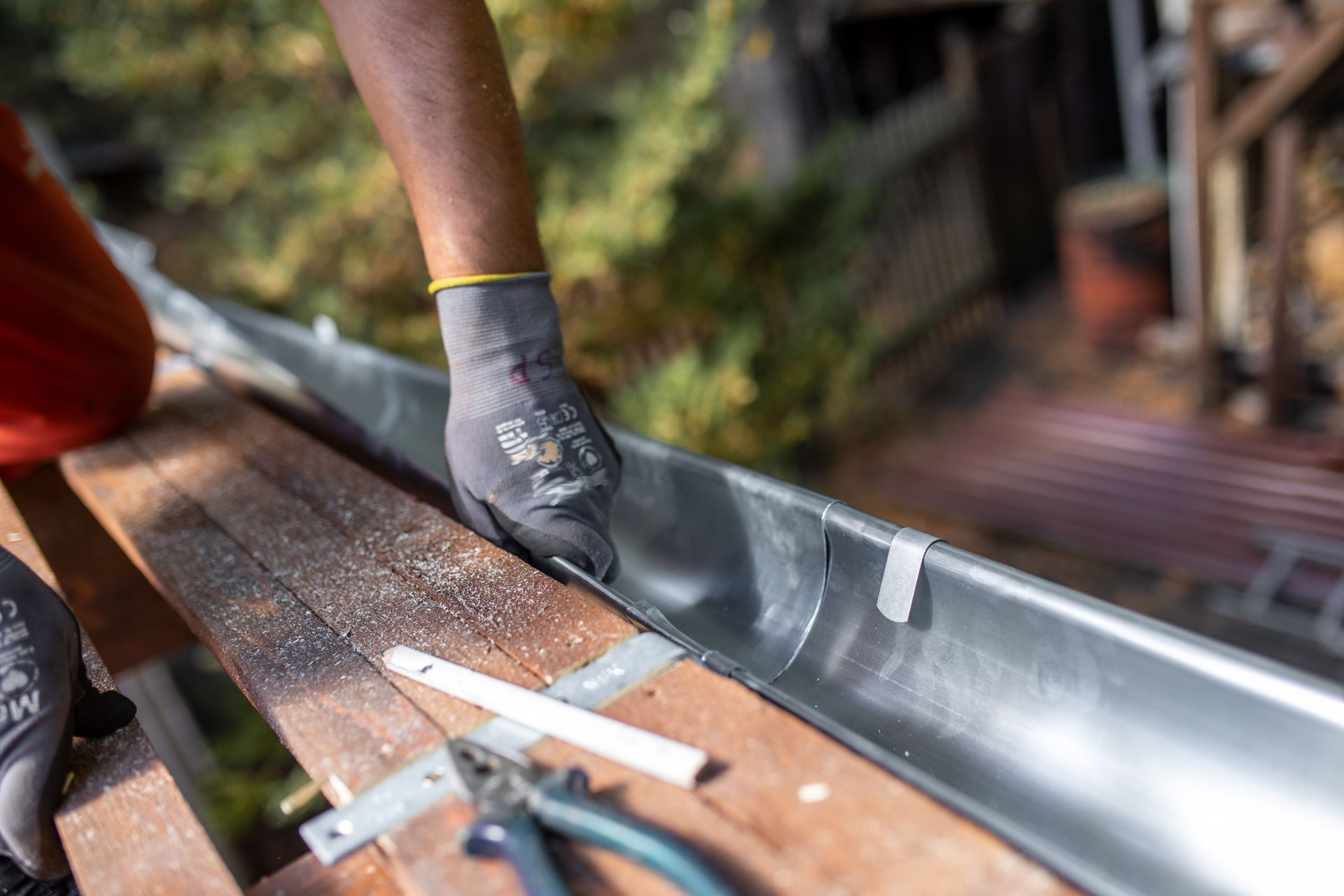 Une personne portant des gants de travail installe une gouttière métallique sur une planche de rive en bois, à l'aide d'outils situés sur l'échafaudage voisin.