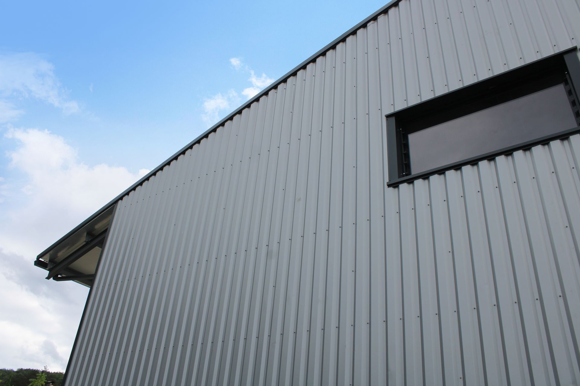 Vue en contre-plongée d'un mur d'immeuble en tôle ondulée grise se détachant sur un ciel bleu partiellement nuageux, avec une fenêtre rectangulaire.
