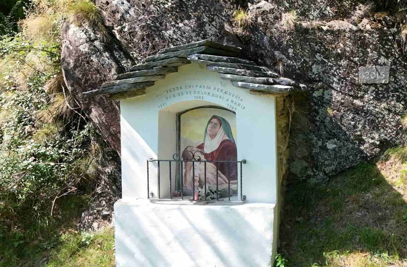 Un piccolo santuario bianco costruito su una parete rocciosa, con un tetto in pietra e una piccola ringhiera in ferro.