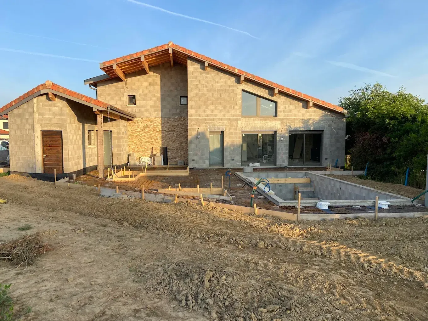 Une maison en pierre de deux étages en construction, avec un espace piscine, par une journée ensoleillée.