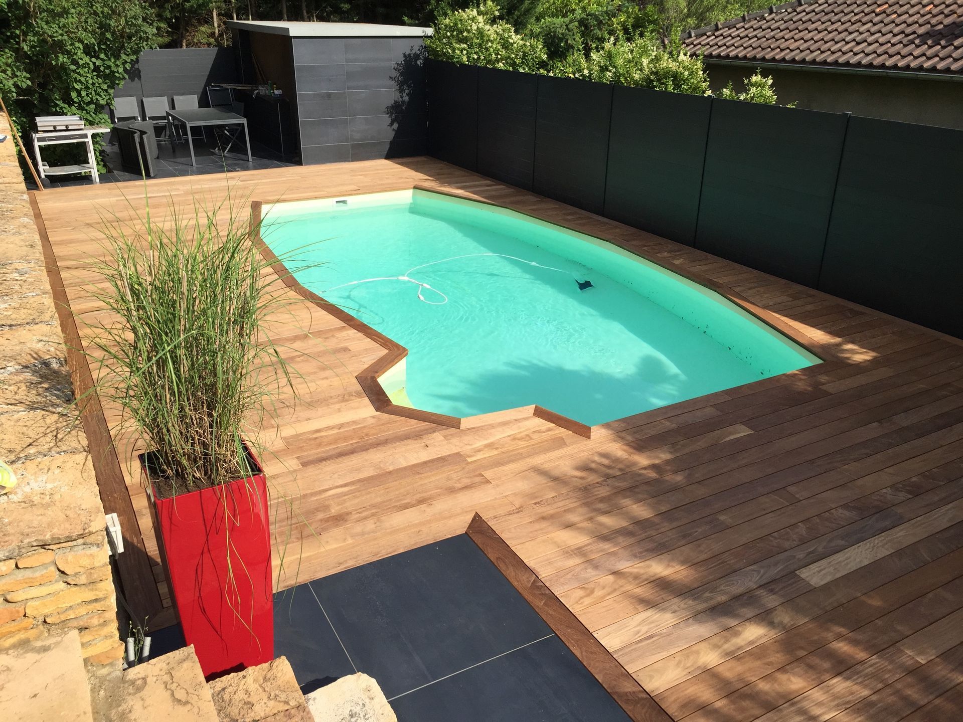 Piscine avec terrasse en bois, clôture foncée et jardinière rouge.