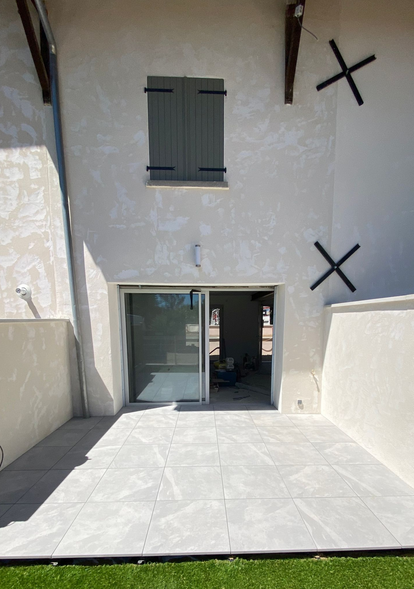 Terrasse arrière avec porte-fenêtre coulissante, carrelage gris, pelouse verte et fenêtre à volets gris.