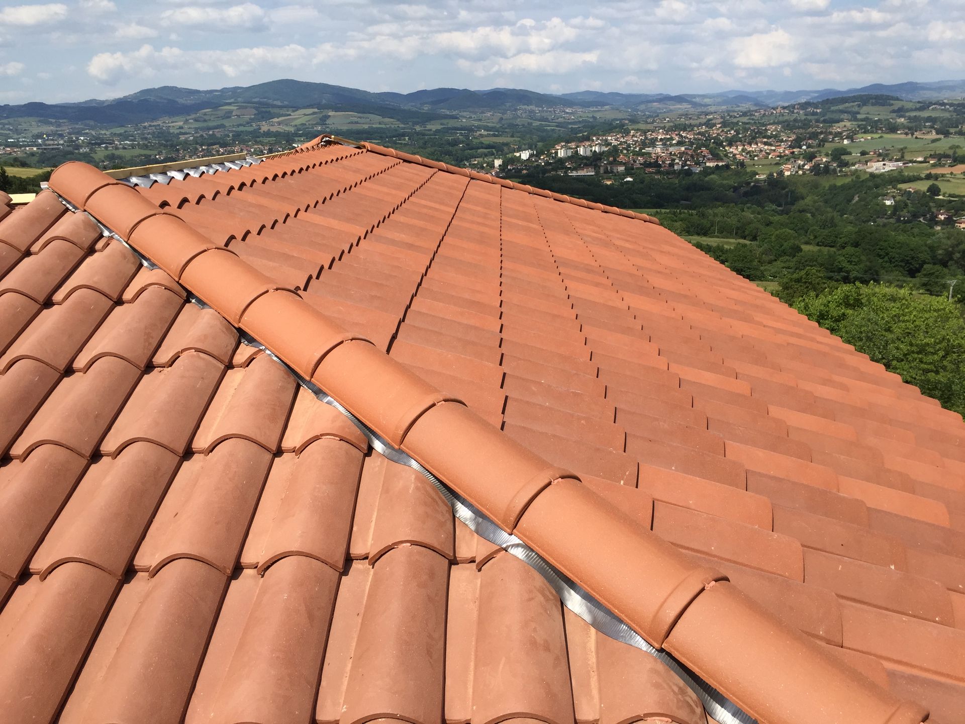 Toit en tuiles de terre cuite rouge avec faîtières et noues, donnant sur une ville et des collines.