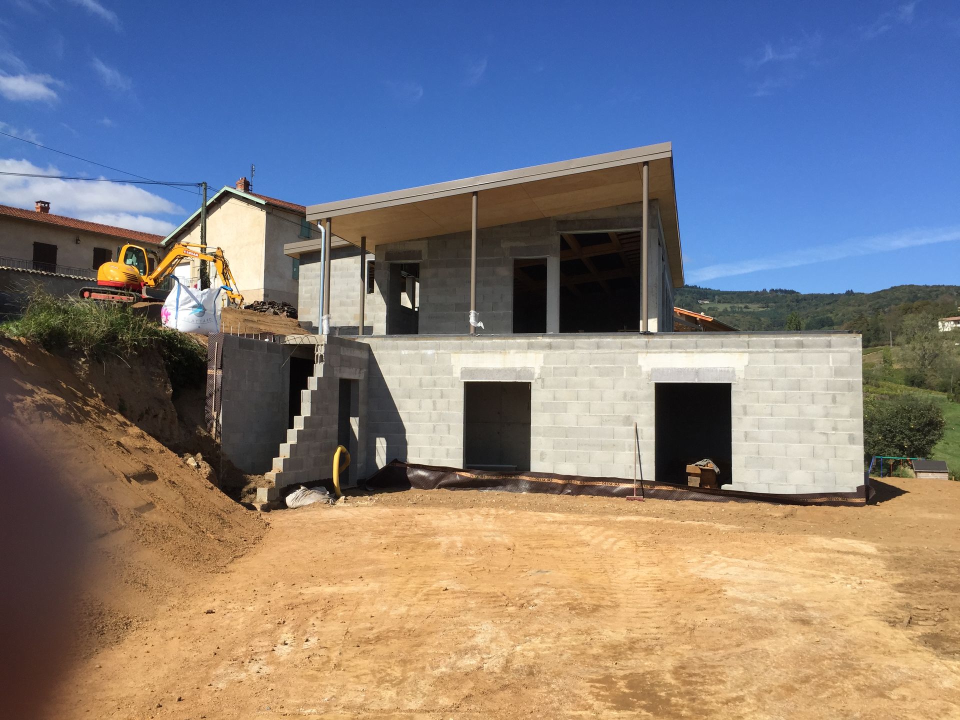 Maison en construction, murs en parpaings gris, poutres apparentes, environnement aux tons naturels, ciel dégagé.
