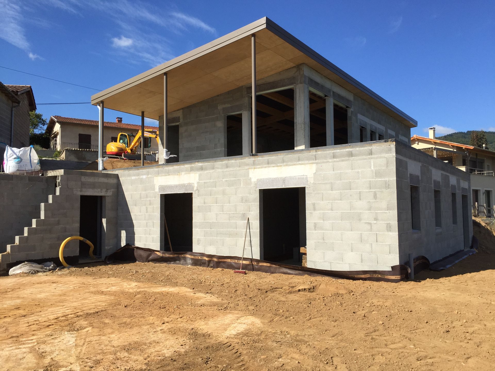 Maison en construction avec des murs en parpaings, un toit plat et une excavatrice en arrière-plan.