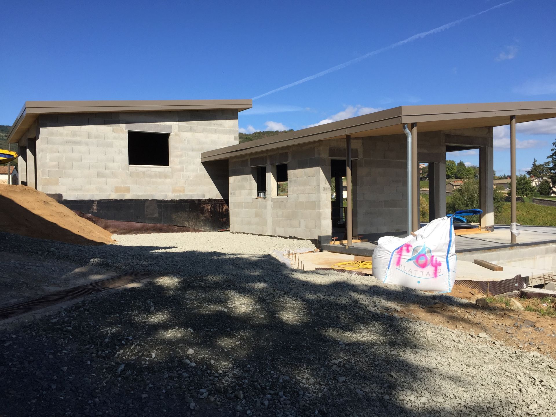 Chantier de construction avec deux bâtiments en parpaings gris sous un ciel bleu. Gravier au premier plan.