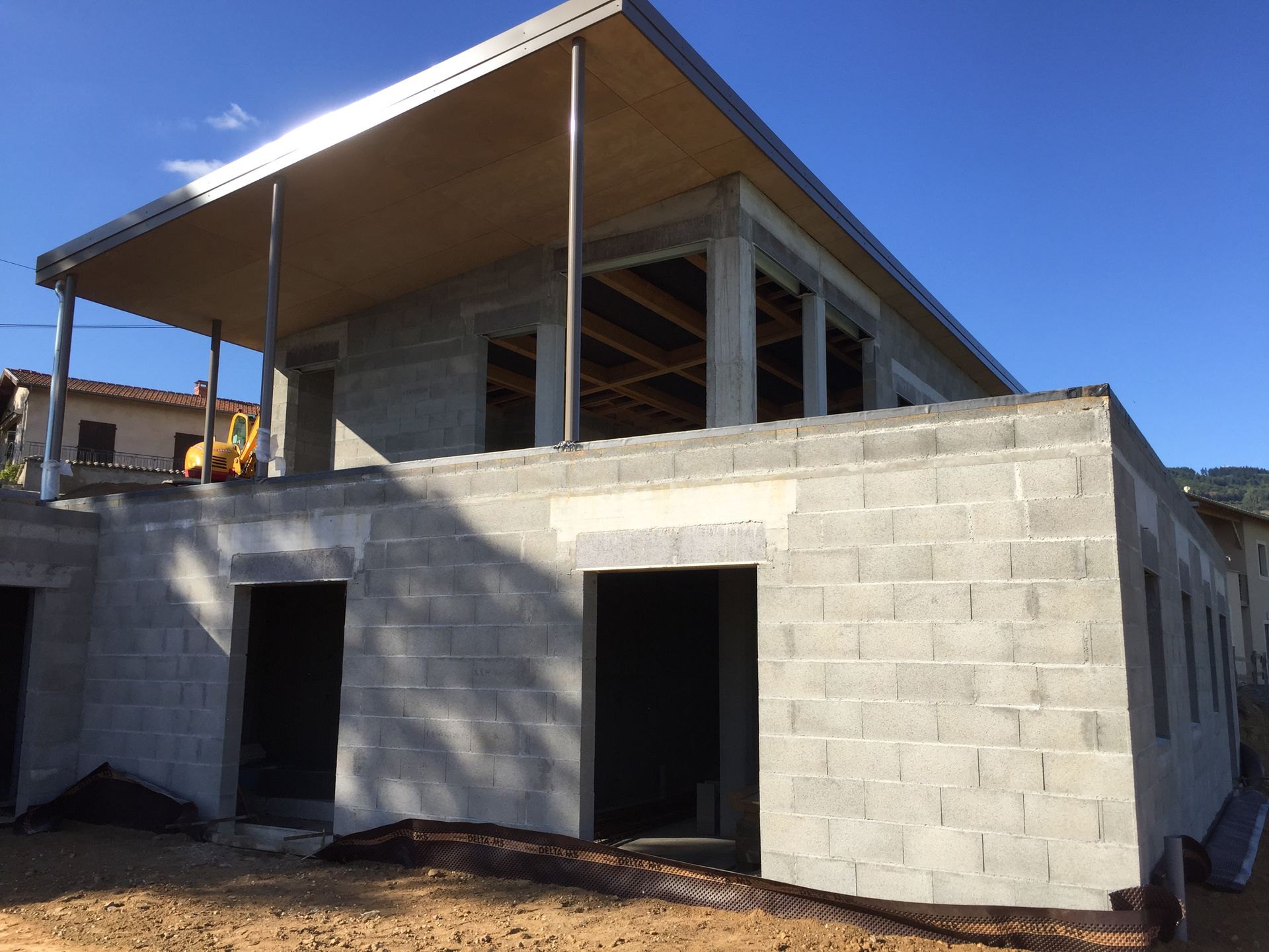 Bâtiment en construction, murs en parpaings, toiture partielle et porche. Ciel dégagé.