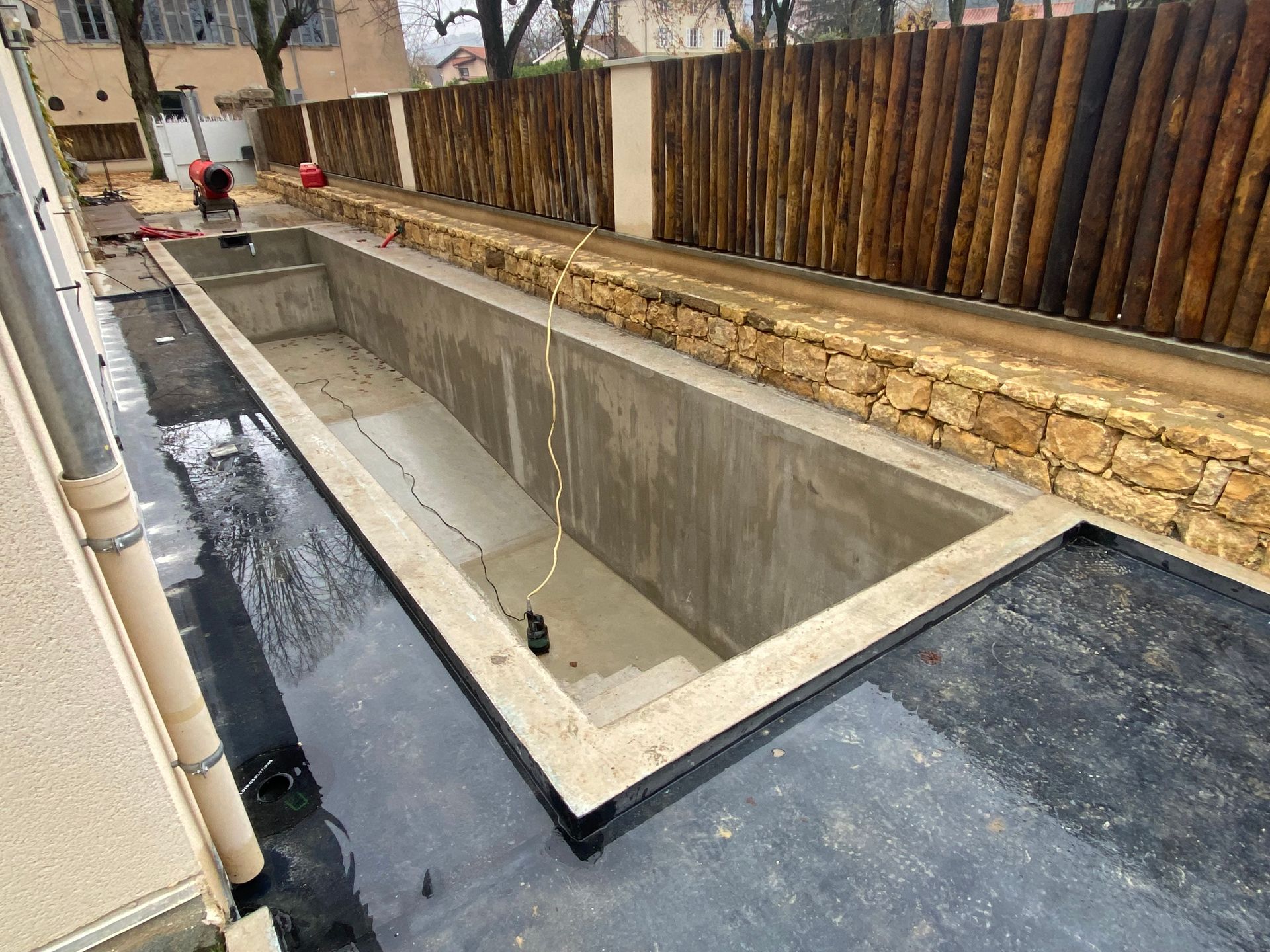 Une piscine en béton en construction, le long d'une clôture en bois et d'un mur en pierre.