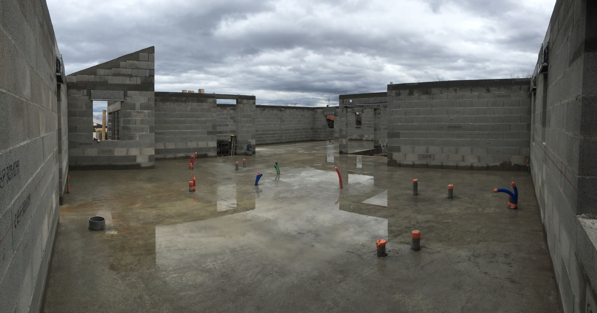 Vue intérieure d'un bâtiment en parpaings en construction, avec un sol mouillé et un ciel nuageux.