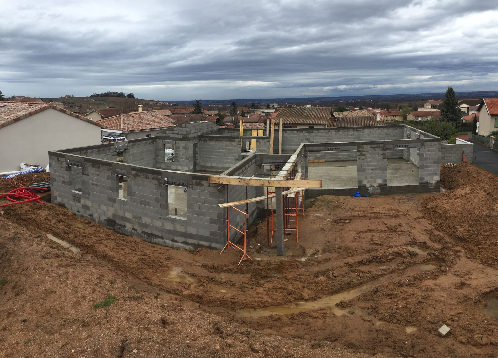Chantier avec un bâtiment en parpaings gris partiellement construit. Terre environnante et ciel couvert.