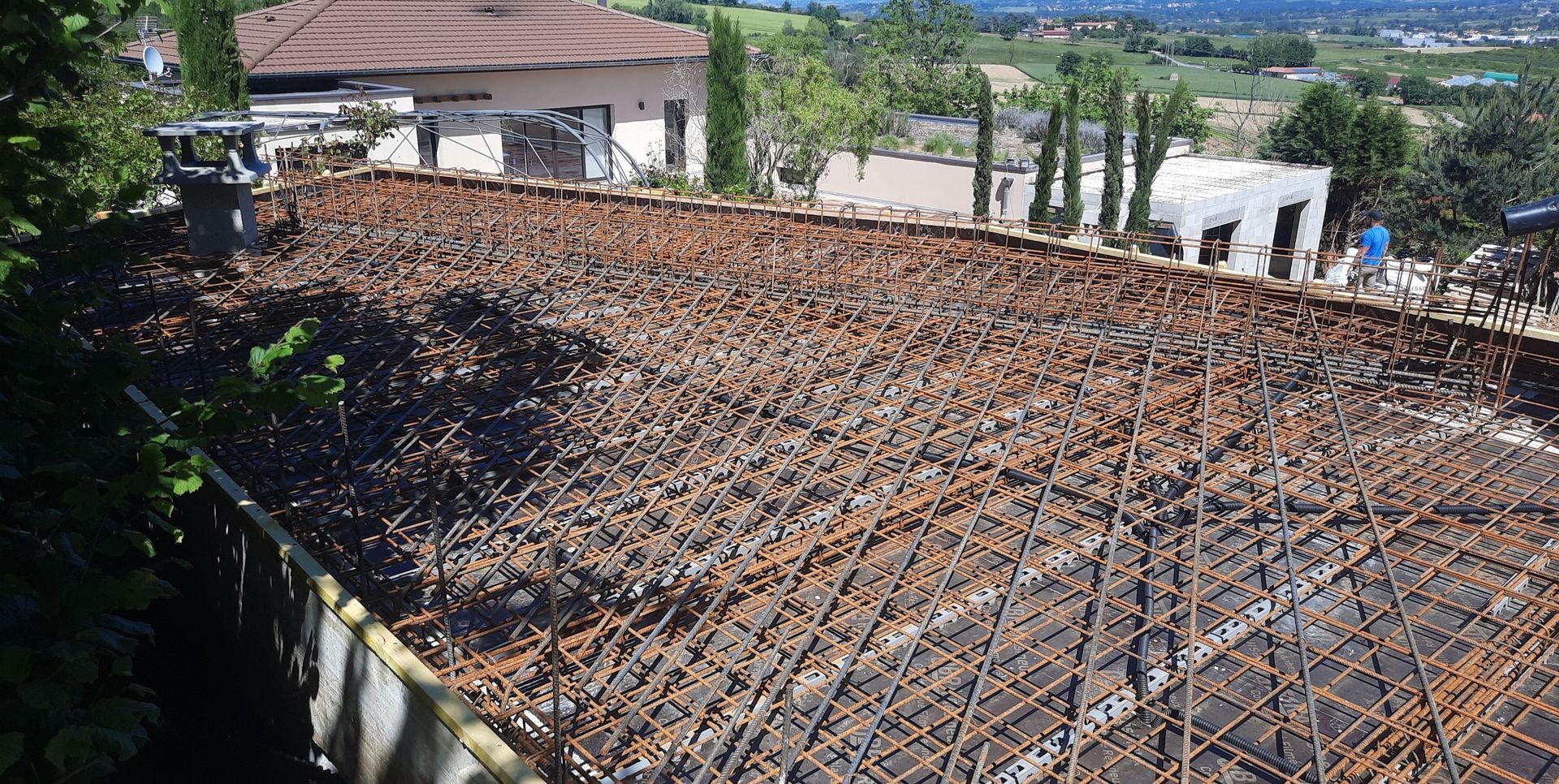 Chantier, treillis d'armature, préparation de la dalle en béton. Bâtiment en arrière-plan, arbres et vue sur le paysage environnant.