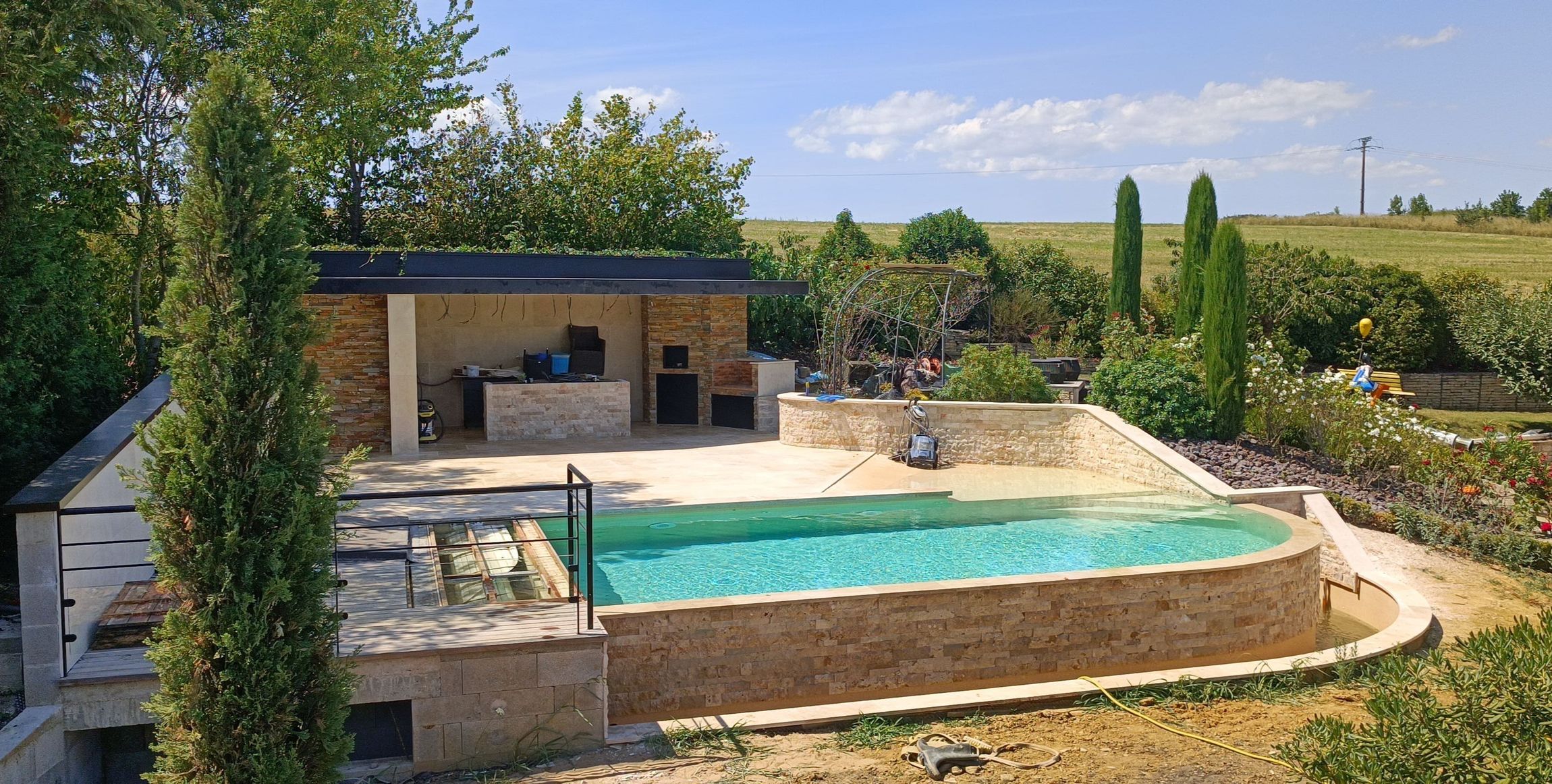 Terrasse avec piscine, éléments en pierre et cuisine ouverte, dans un cadre champêtre.