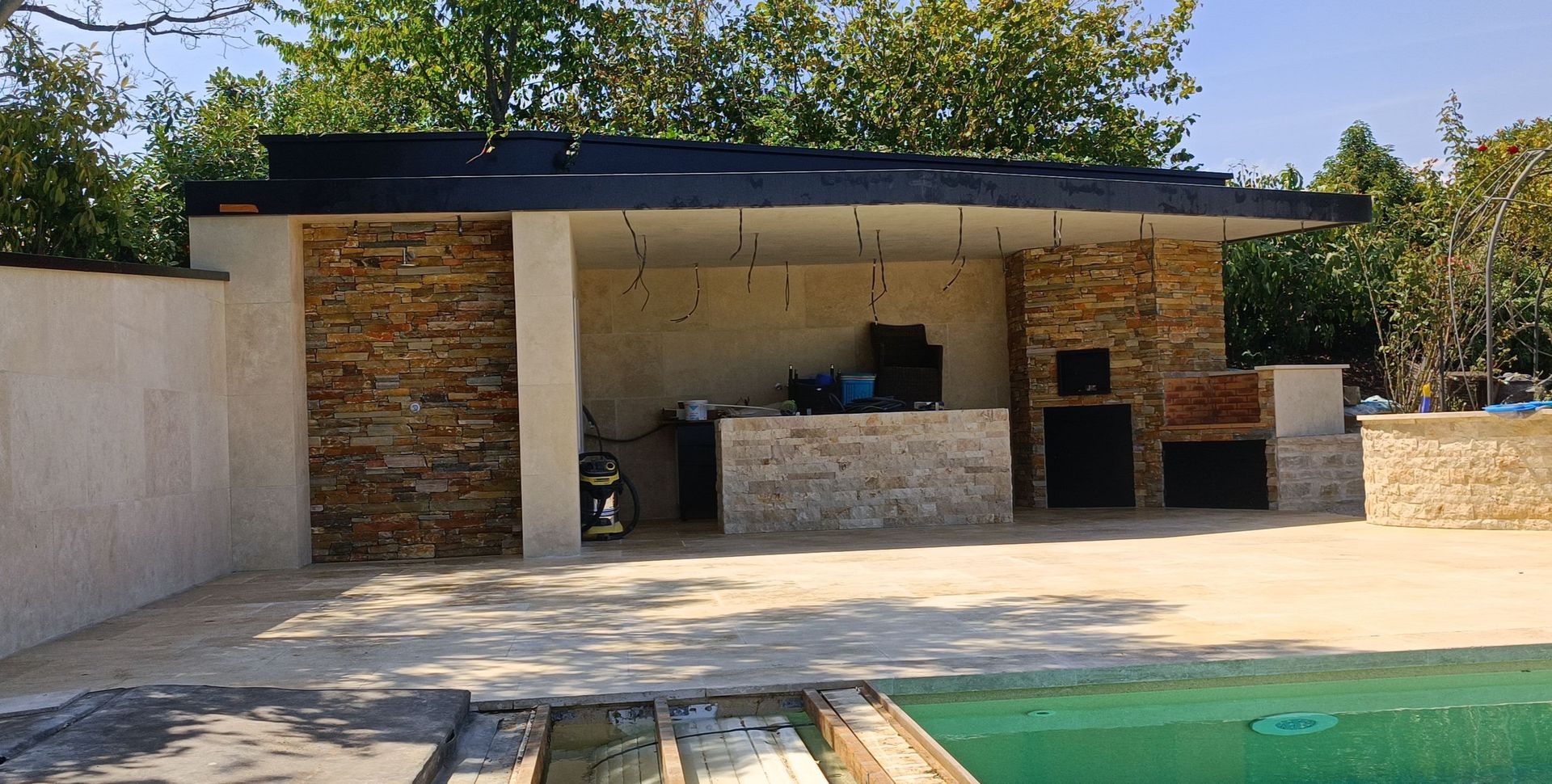 Cuisine extérieure avec éléments en pierre, piscine visible au premier plan, sous un espace couvert.