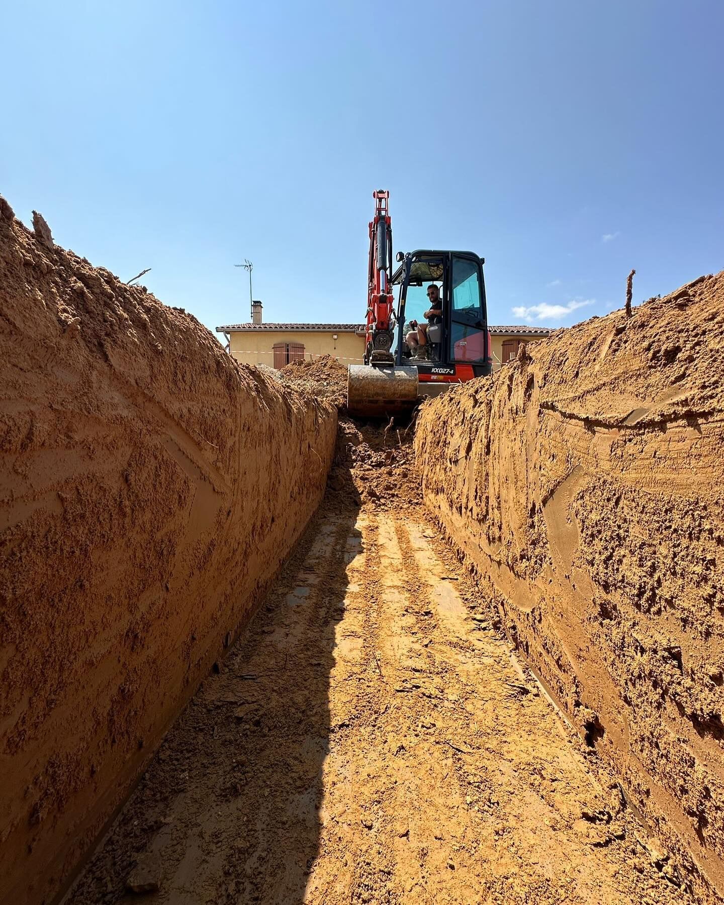 Une excavatrice creuse une longue tranchée dans un sol brun sous un ciel bleu, avec un bâtiment en arrière-plan.