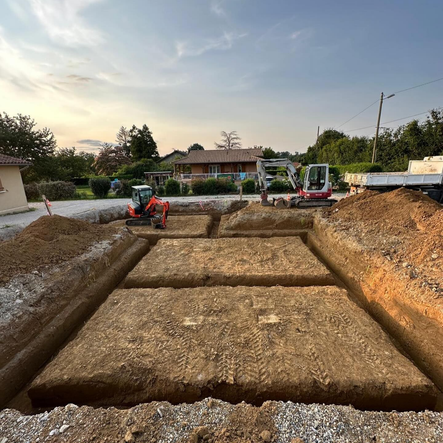 Chantier de construction : Fondations excavées pour un bâtiment, avec des mini-excavatrices et un camion dans une zone résidentielle.