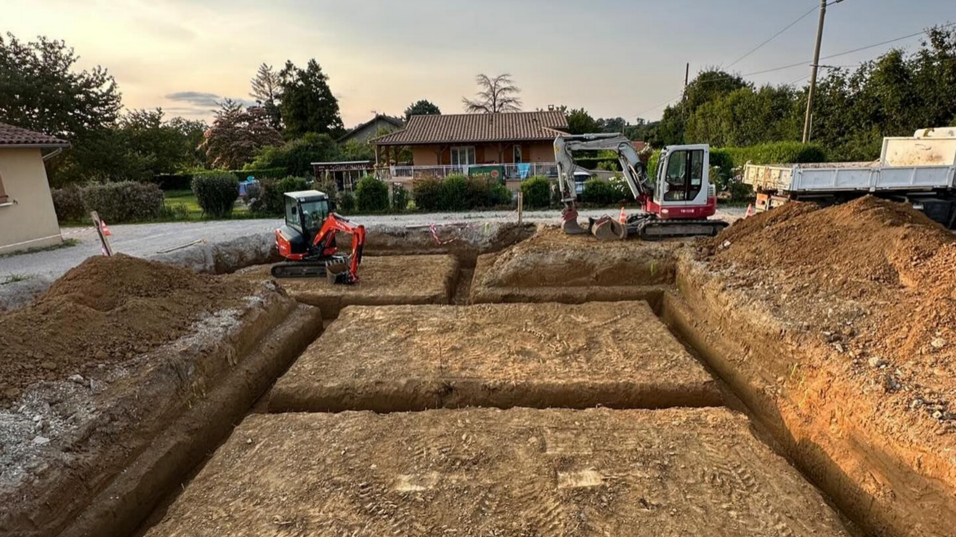 Chantier de fouilles avec des mini-pelles creusant des tranchées rectangulaires pour les fondations ; une maison en arrière-plan.