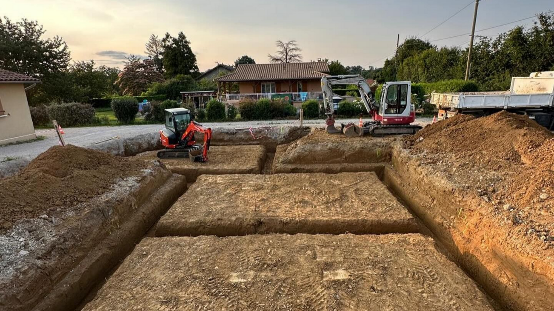 Chantier de construction avec des excavatrices creusant les fondations d'un bâtiment, avec des tas de terre et un arrière-plan résidentiel.