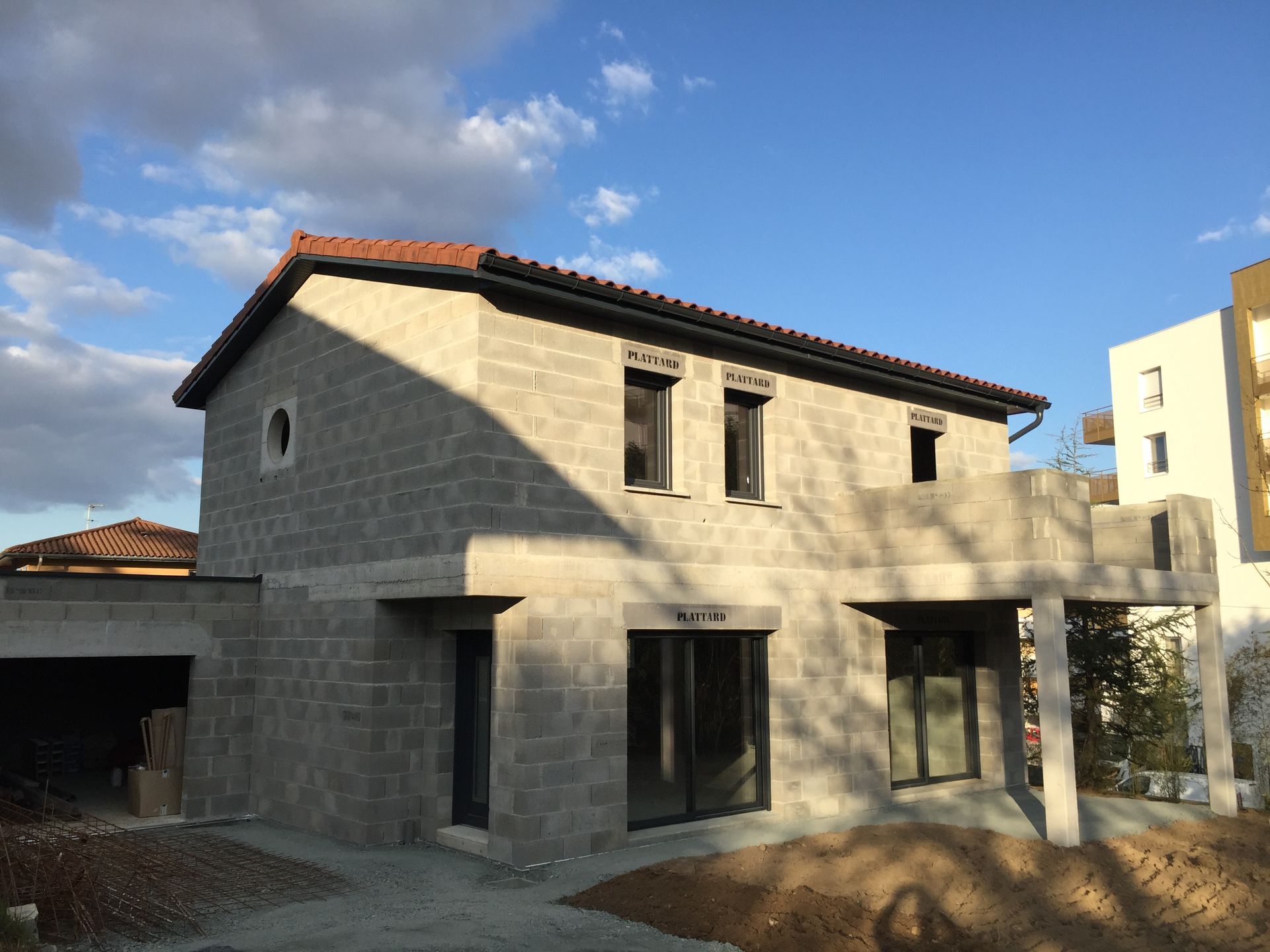 Maison à deux étages en construction, murs en parpaings gris, toit de tuiles rouges, ciel bleu clair.
