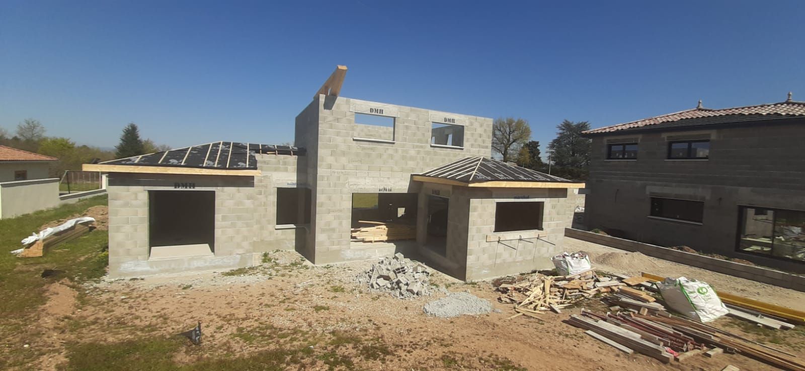 Une maison en construction, avec ses blocs de béton apparents, entourée de matériaux de construction, sous un ciel bleu clair.