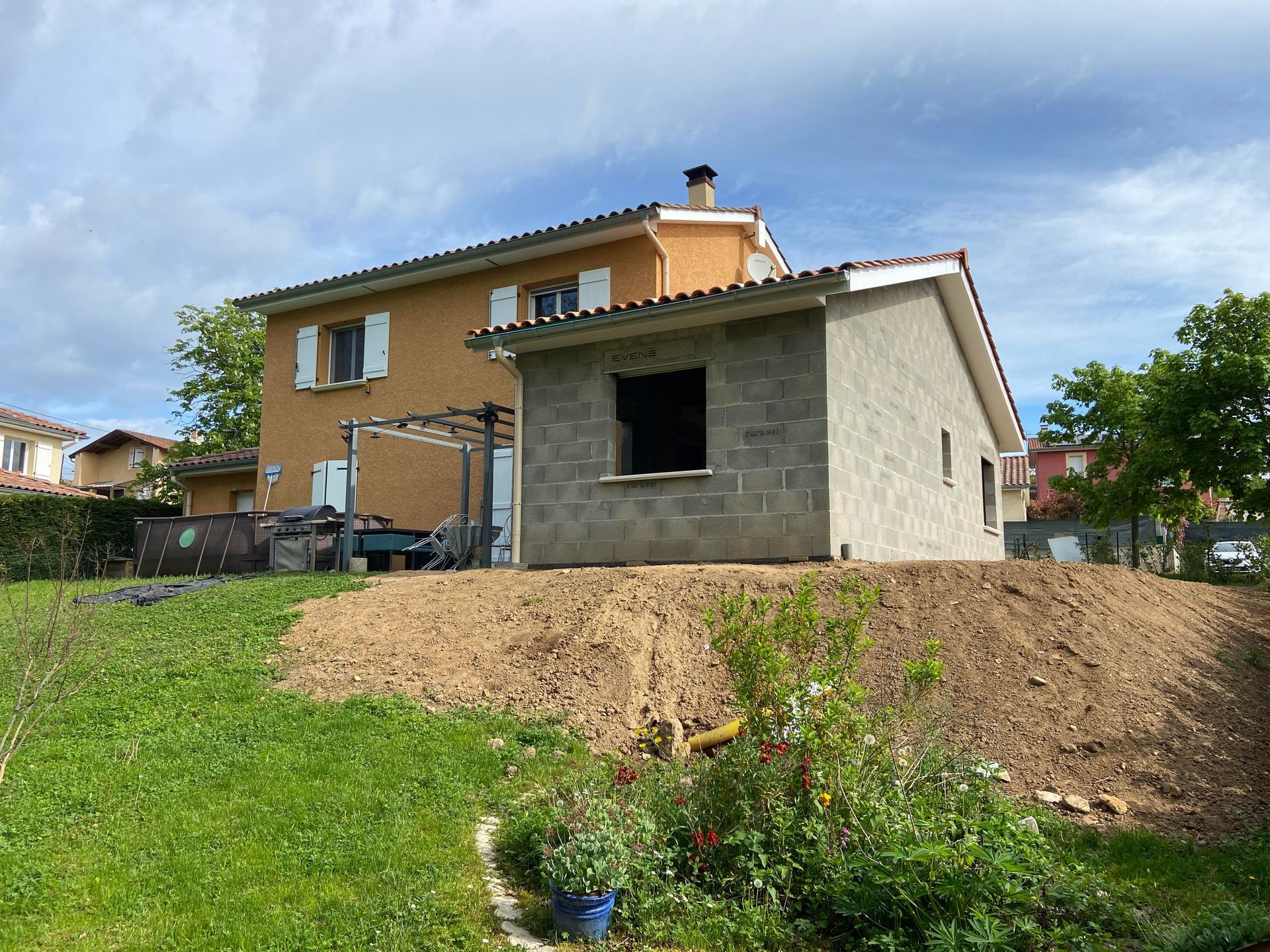 Une maison orange aux volets blancs, un bâtiment en parpaings gris en construction et une cour gazonnée.