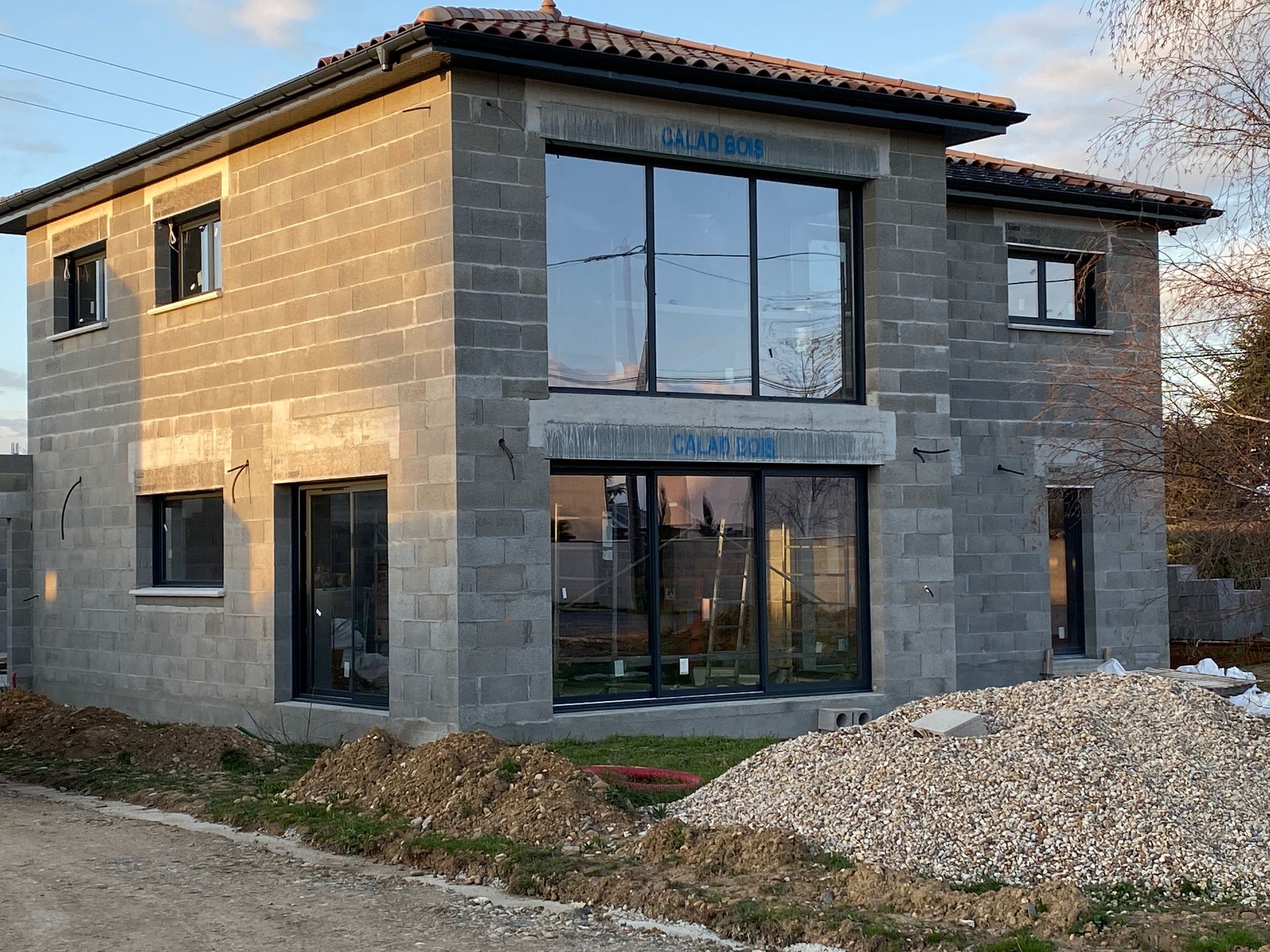 Maison à deux étages en construction, murs en parpaings de béton gris, grandes fenêtres, tas de gravier.