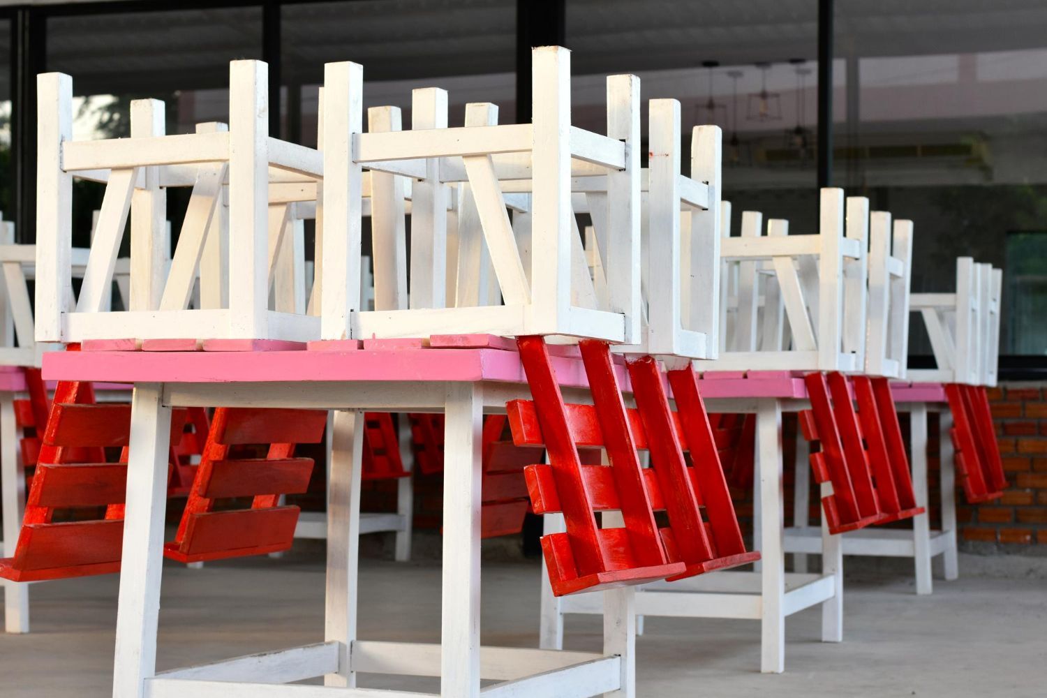 Sillas blancas apiladas sobre mesas rojas, posiblemente después del cierre, al aire libre.
