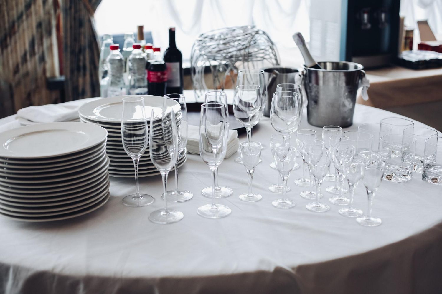 Mesa preparada para un evento con pilas de platos blancos, copas de vino y botellas de vino y agua.
