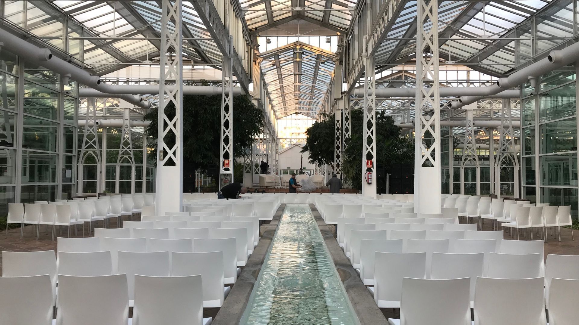 Filas de sillas blancas frente a una fuente de agua en un invernadero de cristal.