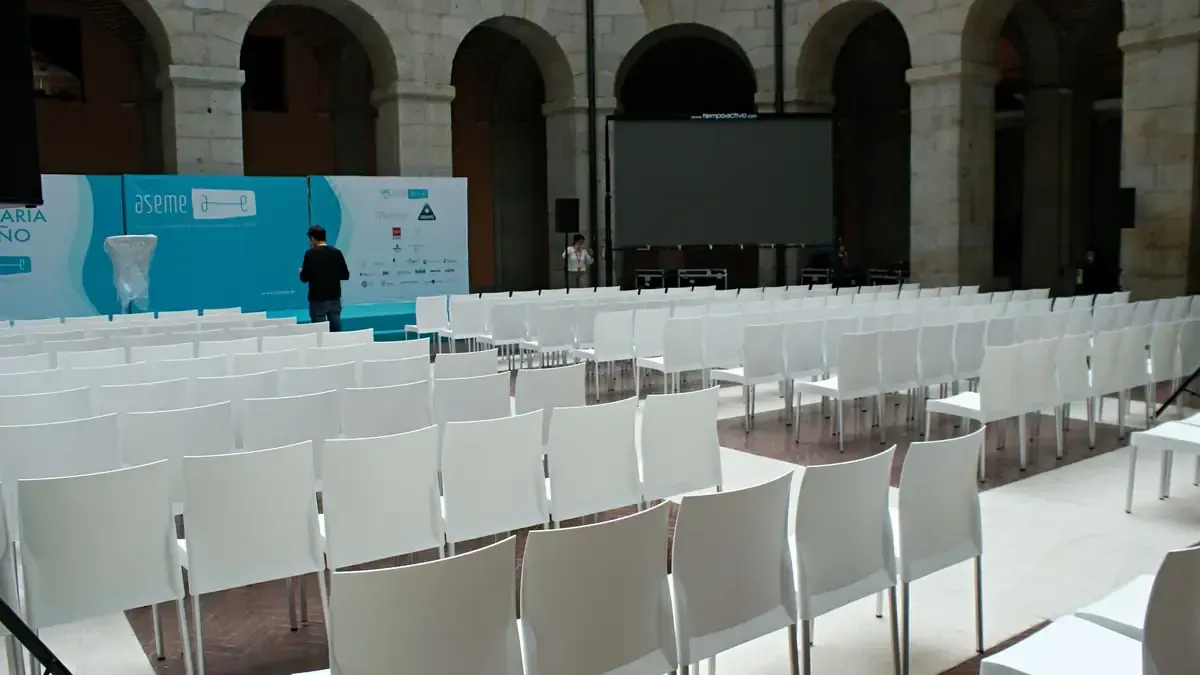 Filas de sillas blancas dan a un escenario con fondo azul y pantalla en una sala arqueada.