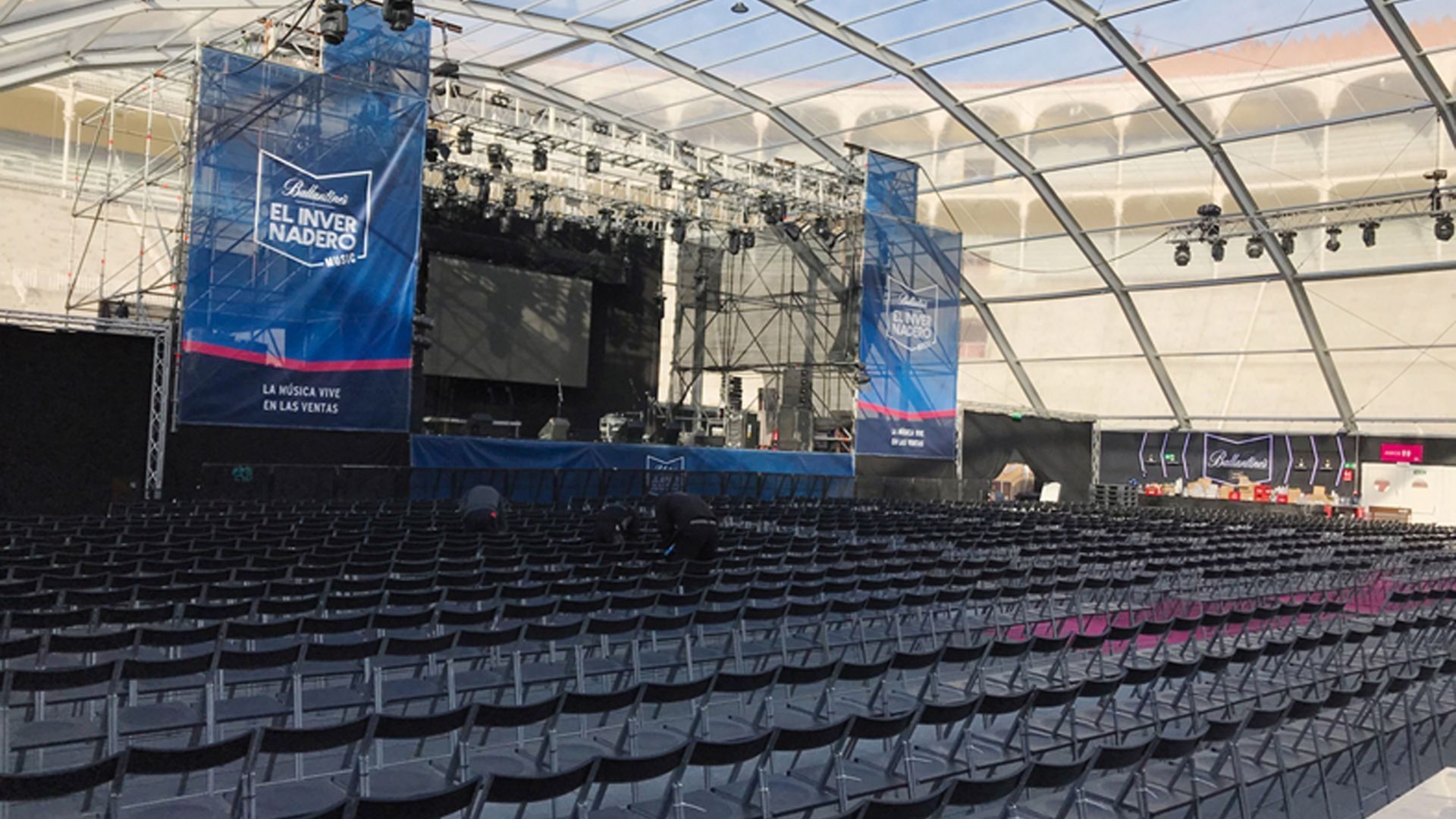 Filas de sillas vacías frente a un escenario bajo una carpa; pancartas cuelgan del escenario.