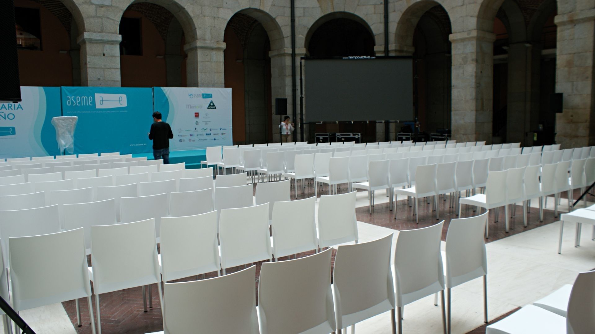 Sillas blancas vacías frente a un escenario con una pantalla en un gran salón con arcos.