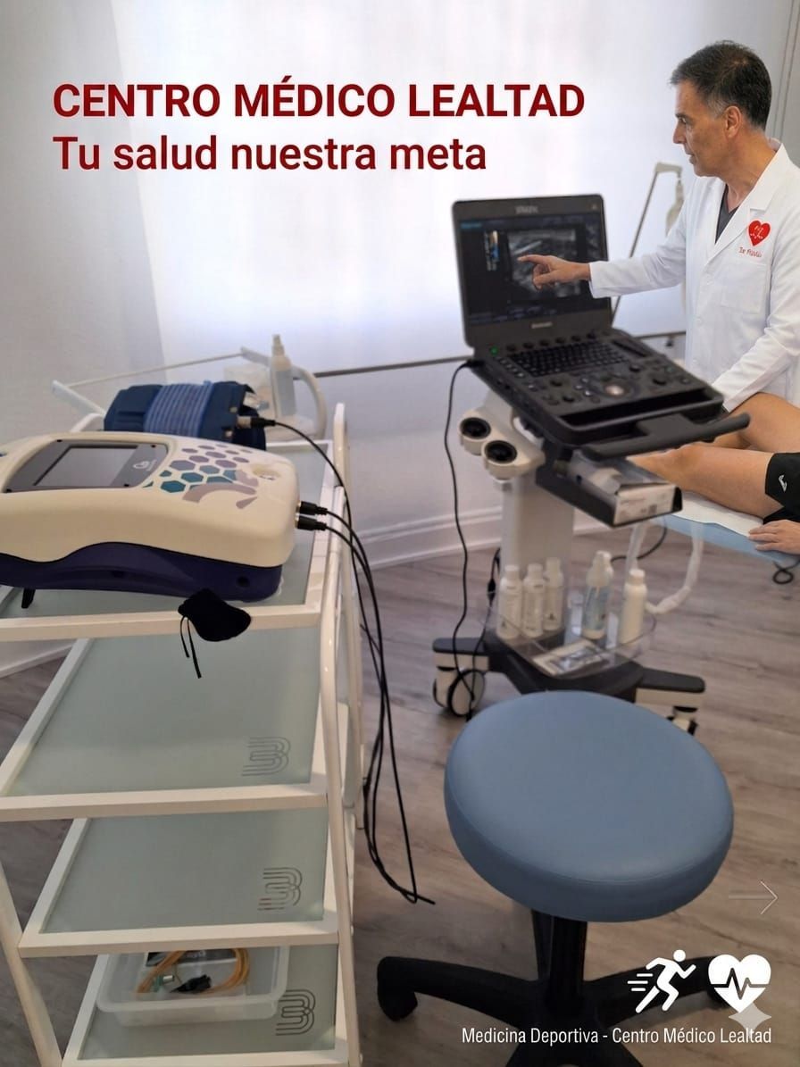 Sala de exploración clínica con ecógrafo, taburete y un médico junto a un ordenador portátil; letrero de “Centro Médico Lealtad”.