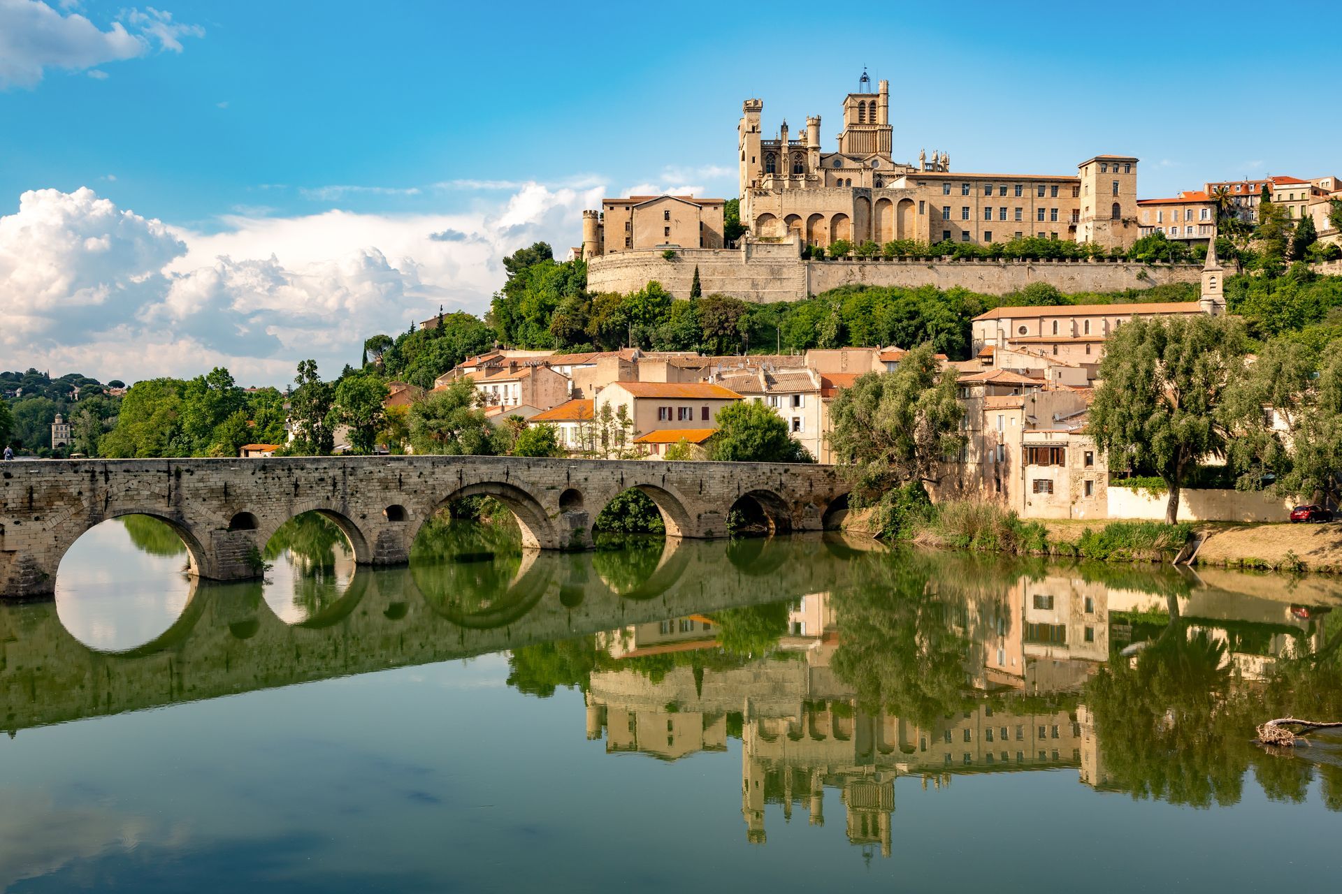 Pont de pierre, bâtiments et cathédrale se reflétant dans l'eau calme sous un ciel bleu à Béziers, France.