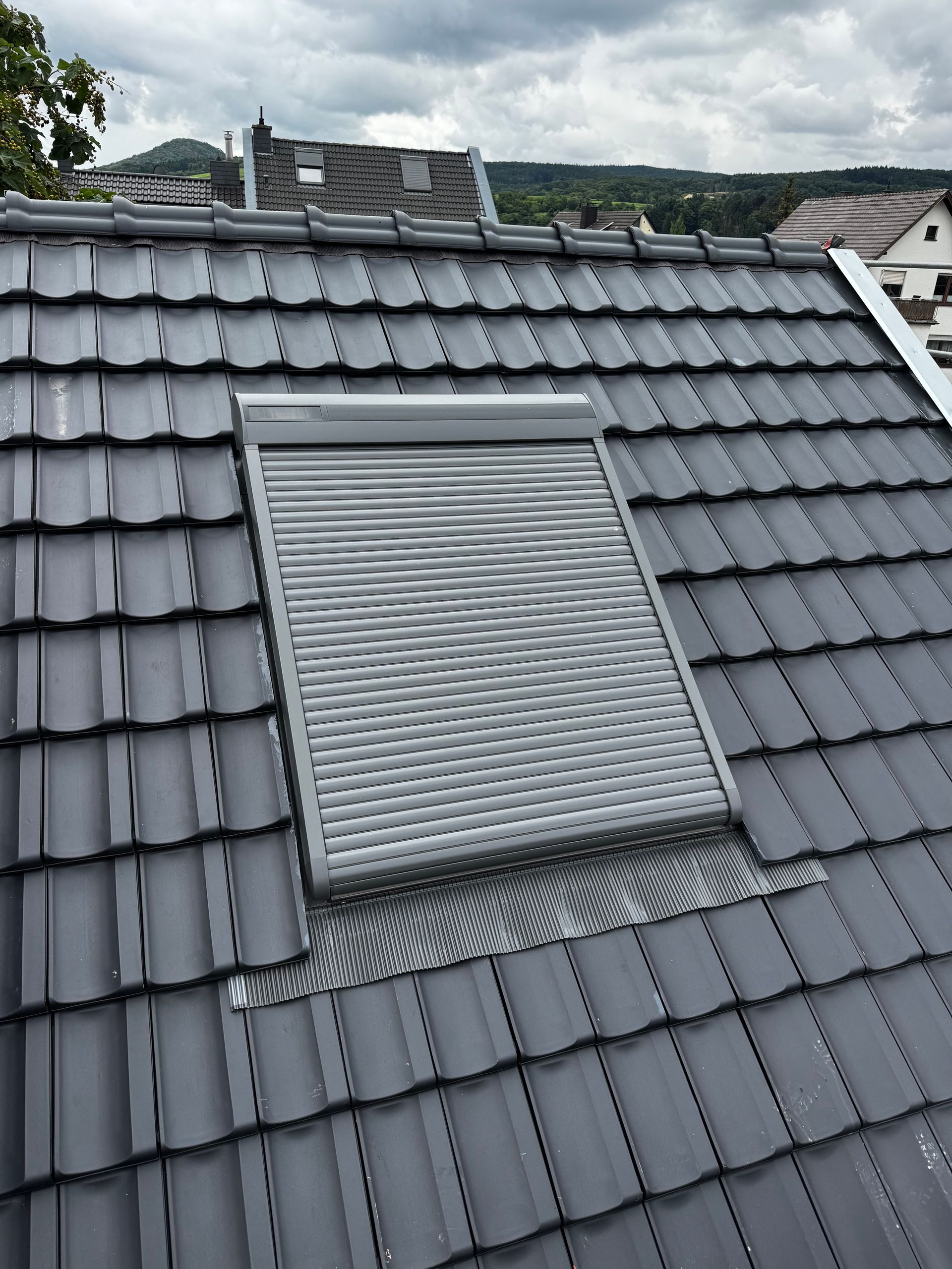 Dach mit Dachfenster und geschlossenem grauen Rollladen auf dunkelgrauen Dachziegeln.