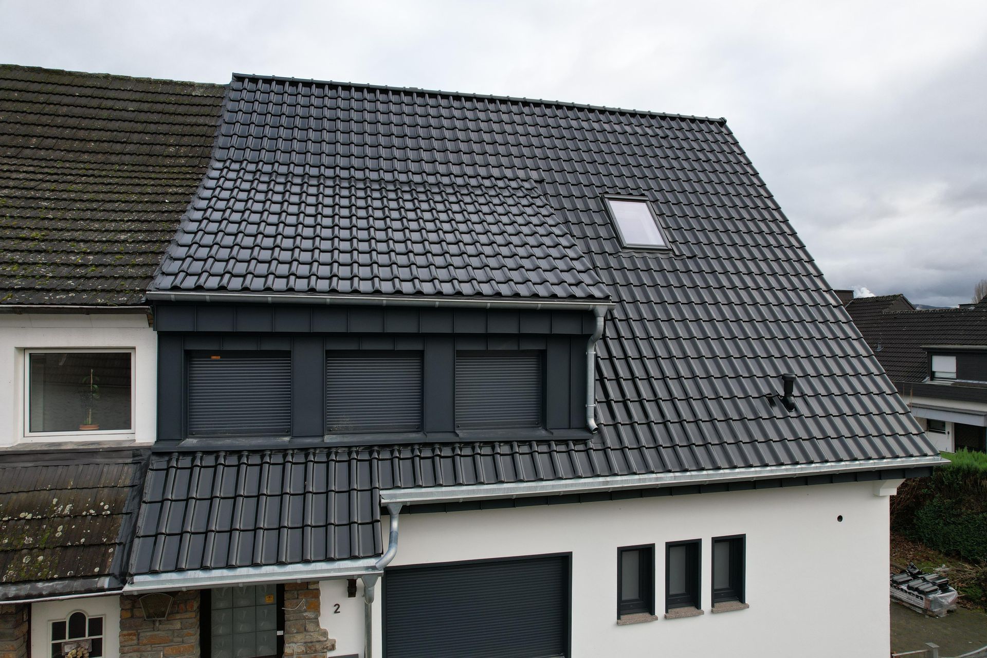 Zwei sich kreuzende Hausdächer mit grauen Schindeln, Dachgauben, Ziegelwänden und einer Satellitenschüssel