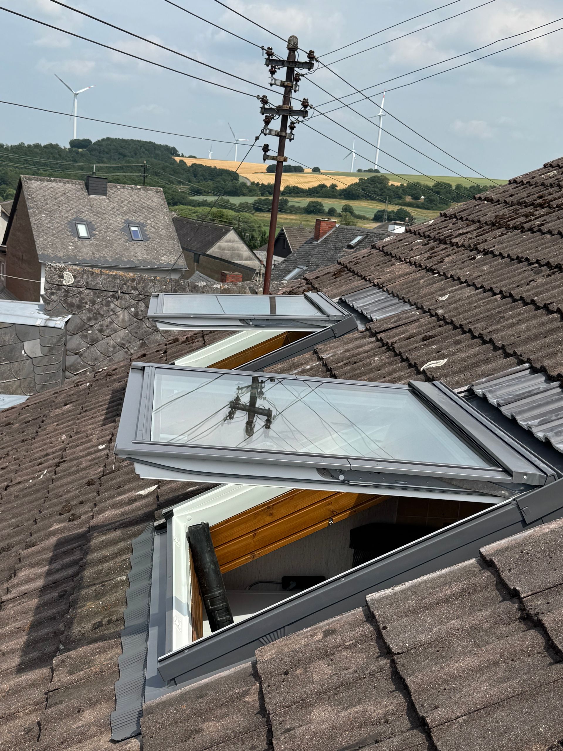Dach mit geöffneten Dachfenstern und sichtbaren Schäden, mit Blick auf ein ländliches Dorf und Stromleitungen.