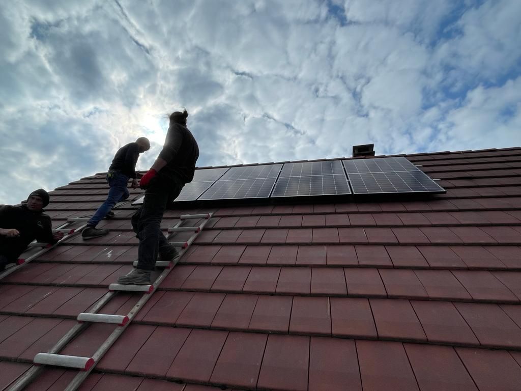 Des ouvriers installent des panneaux solaires sur un toit de tuiles rouges sous un ciel nuageux.