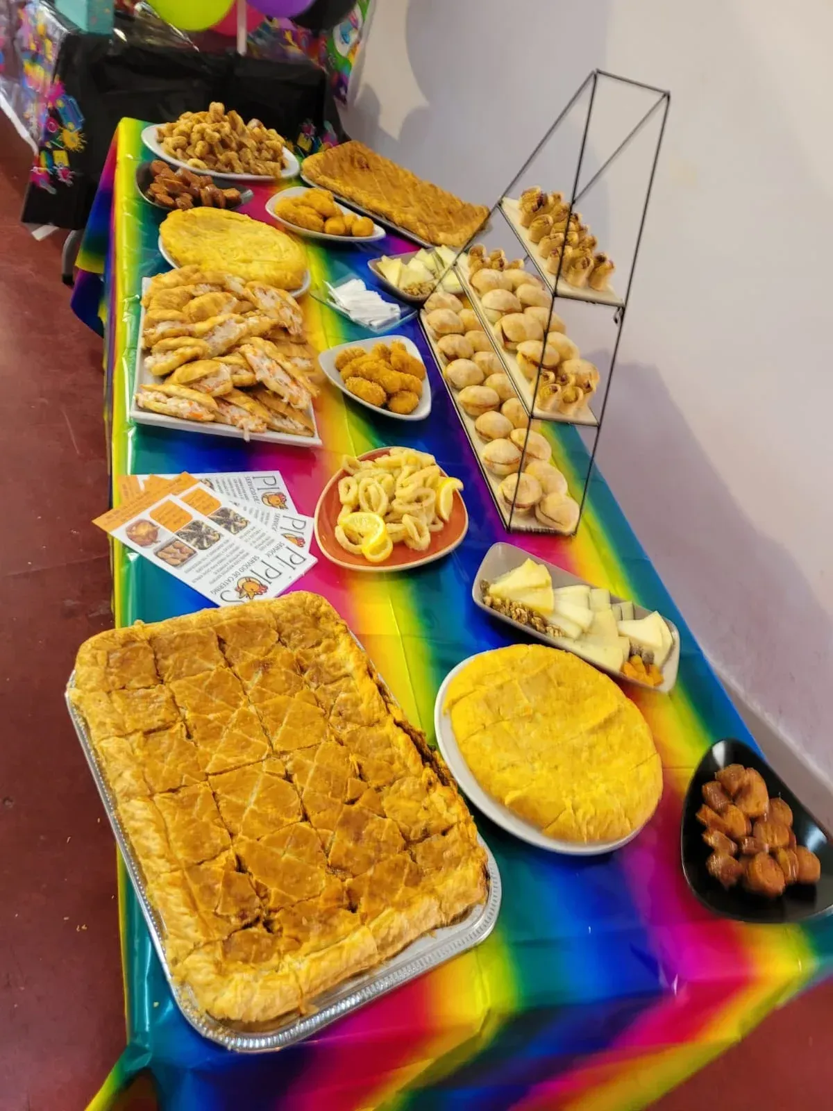 Una mesa repleta de pasteles y bocadillos salados, sobre un mantel de colores del arco iris.