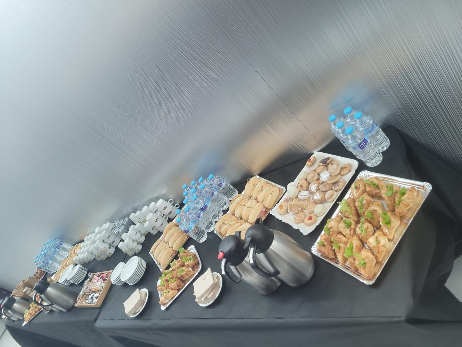 Mesa con aperitivos, bebidas y termos para café: botellas de agua, pasteles y tazas. Mantel negro y fondo metálico.