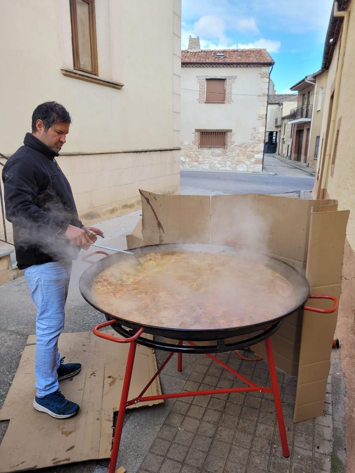 Hombre cocinando paella en una sartén grande al aire libre. Sale vapor. Cortavientos de cartón.