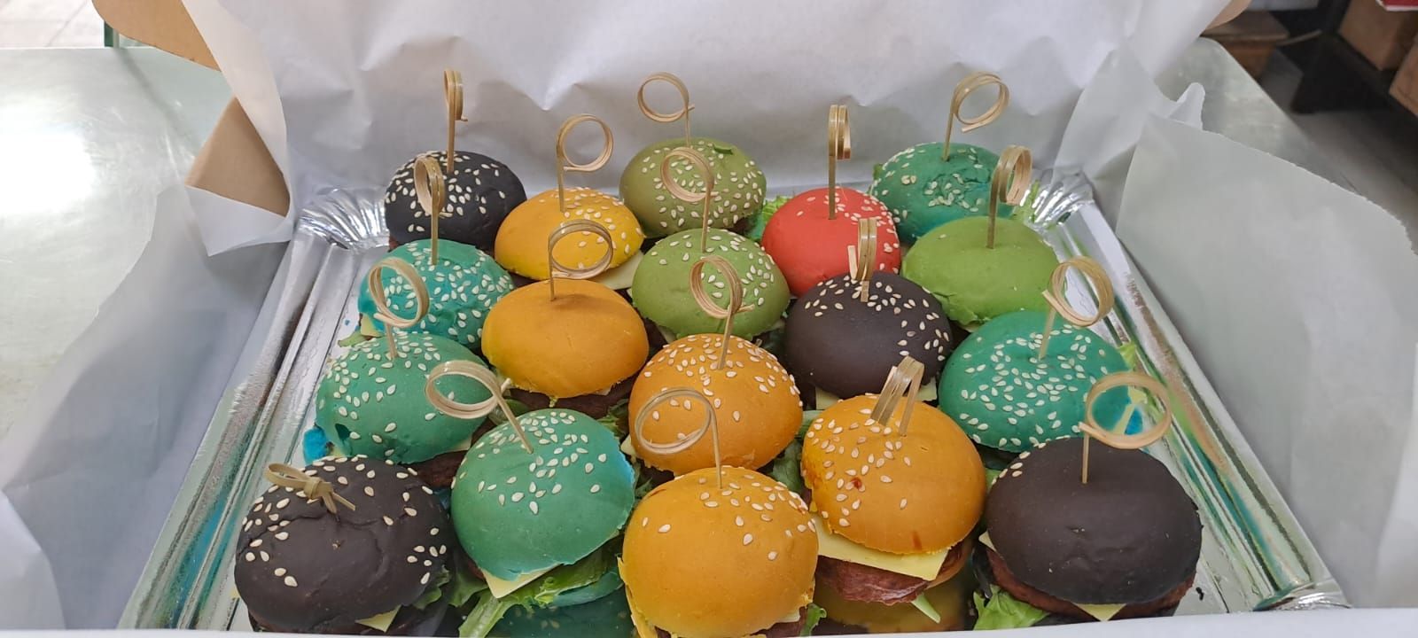 Mini hamburguesas coloridas en una caja, cada una asegurada con un palillo.