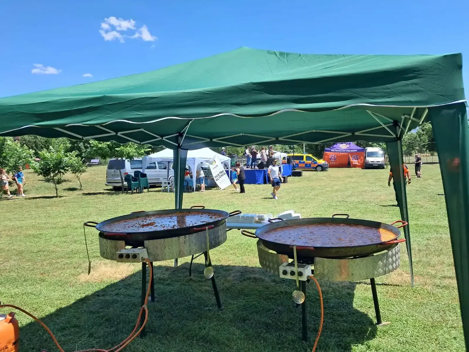 Dos grandes paelleras cocinándose bajo una carpa verde en un evento al aire libre.