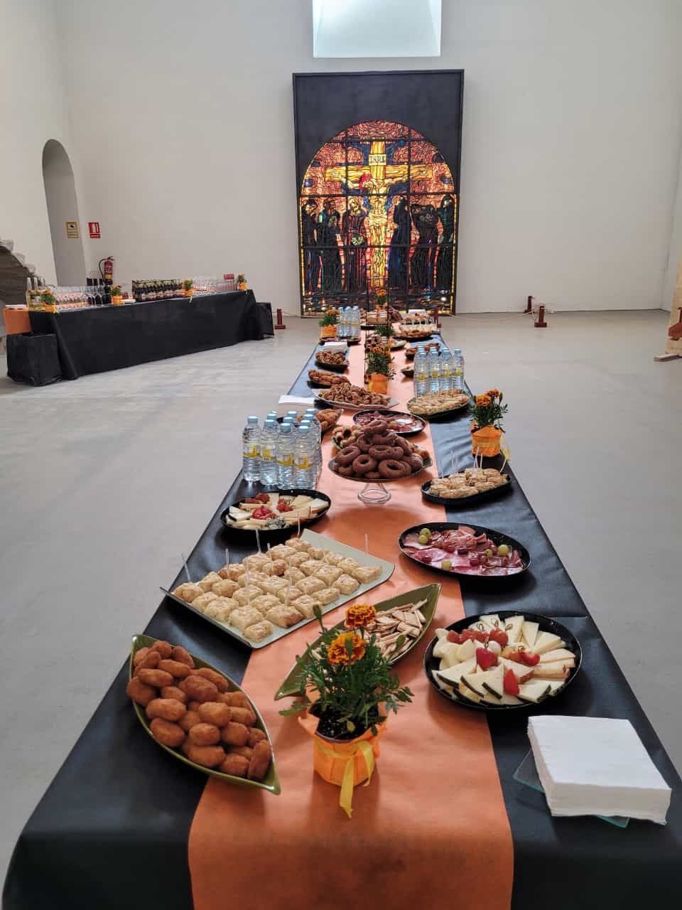 Mesa larga cargada de comida, delante de un crucifijo de vidrieras en una galería.