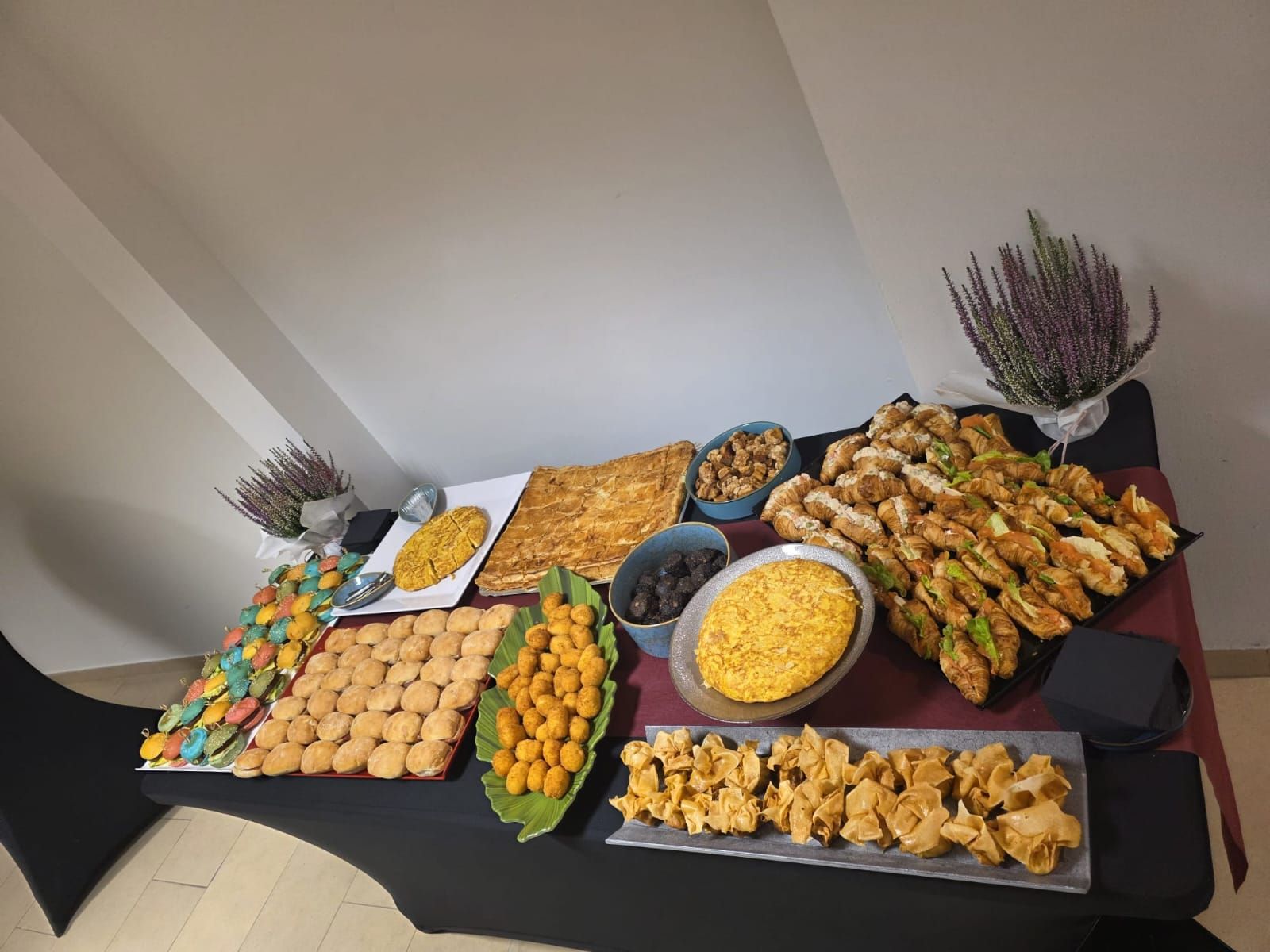 Mesa de buffet con diversos aperitivos: pasteles, sándwiches, frutas y postres. Plantas de brezo decoran el rincón.