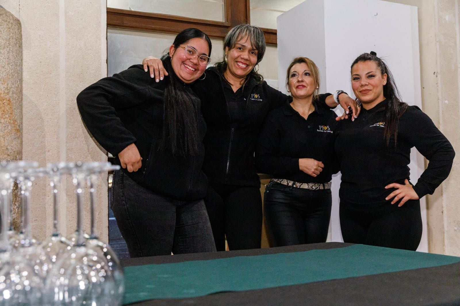 Cuatro personas vestidas con camisa y pantalones negros posan juntas cerca de una mesa y cristalería.